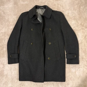 Tung och elegant vinterkappa i mörkgrå – mycket hög kvalitet, helt ny -  Stilren och elegant vinterkappa i mörkgrå färg. Tillverkad i ett tjockt och tungt material som ger fantastisk värme och en lyxig känsla. Dubbelknäppt design med praktiska sidofickor och dekorativa knappar vid ärmsluten för en sofistikerad look.  🧶 Skick: Helt ny (aldrig använd) 🌡️ Egenskaper: Mycket varm, kraftig kvalitet, perfekt för kalla vinterdagar 🎩 Stil: Klassisk / elegant / tidlös  