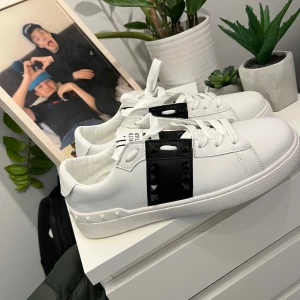 Valentino skor med svarta nitar - Snygga vita sneakers med svart detalj och svarta nitar på sidorna. Skorna har snörning och en platt sula. Märket står tydligt på plösen och designen är riktigt clean med en edgy touch. Perfekta för dig som vill sticka ut lite extra.