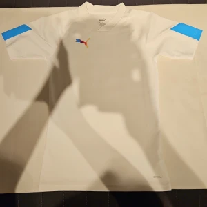 Vit Puma fotbollströja XS - Snygg vit fotbollströja från Puma i storlek XS. Tröjan har korta ärmar med blå detaljer på axlarna och ett färgglatt Puma-logo på bröstet. Materialet är lätt och andas, perfekt för träning eller match. DryCell-teknologi för att hålla dig torr.