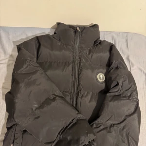 Svart pufferjacka från Trapstar XS - Säljer en svart pufferjacka från Trapstar i storlek XS. Jackan har hög krage, dragkedja framtill och Trapstar-logga på bröstet samt stor broderad Trapstar-text på ryggen. Perfekt för kalla dagar och riktigt street i stilen. Använd 3-4 gånger 