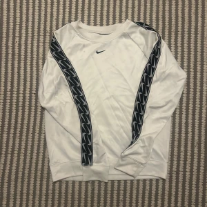 Vit Nike sweatshirt med svarta detaljer XS - Vit långärmad sweatshirt från Nike i storlek XS. Tröjan har svarta mönstrade band längs ärmarna och en liten svart Nike-logga på bröstet. Tillverkad i mjuk polyester med klassisk passform och ribbade muddar vid ärmslut och nederkant.