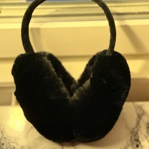 Svarta fluffiga öronmuffar - Snygga svarta öronmuffar med mjuk och fluffig pälsimitation. Perfekt för kalla vinterdagar när du vill hålla öronen varma och samtidigt se cool ut. Bågen är klädd i samma svarta material för en enhetlig look.