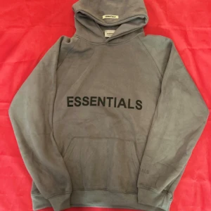 Grå Essentials hoodie från Fear of God - Säljer en grå hoodie från Fear of God Essentials med stor svart logga på bröstet och mindre tryck på ärmen. Klassisk känguruficka, huva och relaxed fit. Perfekt för dig som gillar streetwear och vill ha en clean look.