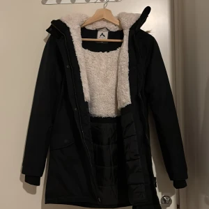 Svart parkasjacka från McKinley, 158 - Svart parkasjacka från McKinley med mysigt vitt teddyfoder och huva med fuskpäls. Jackan har dragkedja, knappar och reflexdetalj på huvan. Perfekt för kalla dagar med sin långa modell och fodrade insida.