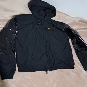 Svart vindjacka från Lyle & Scott - Svart vindjacka från Lyle & Scott med huva och hel dragkedja. Jackan har glansiga svarta ärmar i kontrast mot den matta kroppen, samt en gul logga på bröstet. Tillverkad av återvunnen nylon och har snörning i huvan för extra stil.