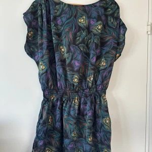 Färgglad kortklänning med påfågelfjädrar - Unik kortklänning med färgstarkt påfågelfjädermönster i blått, lila, gult och turkost på svart botten. Klänningen har resår i midjan, korta ärmar och knapp i nacken. Materialet känns lätt och svalt, perfekt för varma dagar. Snygg och iögonfallande design!