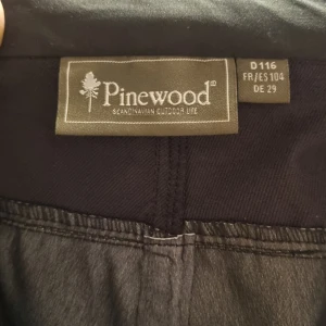 Aldrig använda! Svarta friluftsbyxor från Pinewood - Svarta friluftsbyxor från Pinewood med flera praktiska fickor och dragkedjor. Byxorna har förstärkta partier på knän och bak, samt bälteshällor och knappstängning. Perfekta för utomhusaktiviteter och äventyr.