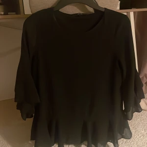 Svart blus med volang från Vero Moda - Svart blus från Vero Moda i storlek XS. Blusen har trekvartsärm och volangdetaljer vid ärmslut och nederkant. Den är tillverkad i ett lätt och flödande material som ger en snygg siluett. Perfekt för dig som gillar stilrena och feminina plagg.