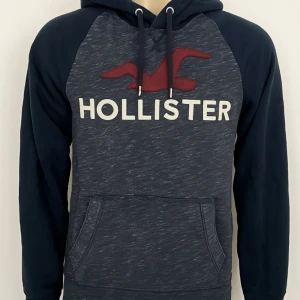 Mörkblå hoodie från Hollister - Snygg mörkblå hoodie från Hollister med stor logga och röd fågel framtill. Tröjan har huva med snörning och en stor magficka. Mjuk insida i fleece och långa ärmar. Perfekt för chill dagar eller när du vill ha en casual look.