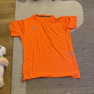 Orange t-shirt från Under Armour - Säljer en snygg orange t-shirt från Under Armour med korta ärmar och rund hals. Perfekt för träning med lätt och mjukt material i polyester. Loggan syns både på bröstet och nedtill på sidan. Skön passform och sportig look!