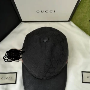 Svart Gucci keps med GG-mönster - Snygg svart keps från Gucci med diskret GG-monogram över hela kepsen och klassisk grön-röd rand på sidan. Kepsen har böjd skärm och knapp på toppen, tillverkad i bomullsmaterial. Perfekt för dig som vill ha en lyxig streetstyle-accessoar.