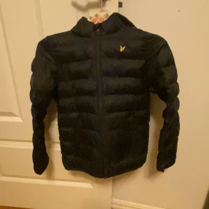 Svart pufferjacka Lyle & Scott 10-11 år - Svart pufferjacka från Lyle & Scott med gul logga på bröstet. Jackan har huva, dragkedja framtill och quiltad design. Passar perfekt för kyligare dagar och har en klassisk look som funkar till det mesta.