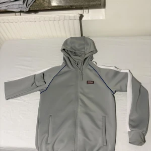 McKenzie hoodie  - Snygg grå hoodie från McKenzie med vita ränder längs ärmarna och diskreta blå sömmar. Tröjan har huva med snörning, dragkedja framtill och två fickor. Perfekt för chill eller träning, stilren och sportig look.