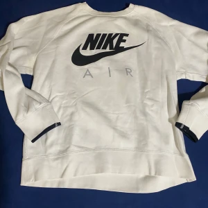 Vit sweatshirt från Nike Air - Snygg vit sweatshirt från Nike Air med stor svart logga framtill. Tröjan har långärmade raglanärmar och ribbade muddar med svart detalj. Mjuk och skön bomullskvalitet, perfekt för chill eller streetwear. Oversized passform för en avslappnad vibe.