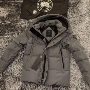 Grå dunjacka från Canada Goose - Säljer en grå dunjacka från Canada Goose med huva och svarta detaljer. Jackan har dragkedja, knappar och två stora fickor framtill. Klassisk patch på ärmen och en clean, stilren look som passar perfekt när det är kallt ute.