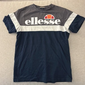 Ellesse t-shirt med logga, navy/grå S - Snygg t-shirt från Ellesse i storlek S med stor logga framtill. Färgerna är mörkblå, grå och vit med en bred vit rand över bröstet och ärmarna. Klassisk rund hals och korta ärmar. Materialet är mjuk bomull, perfekt för en chill look.