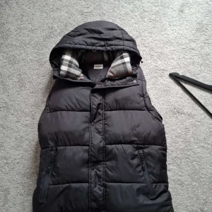 Svart dunväst från Burberry - Svart dunväst från Burberry med huva och klassiskt rutigt foder i grått, vitt och rött. Västen har två fickor med dragkedja och en stor Burberry London England-patch på sidan. Perfekt för kalla dagar och har en quiltad look.