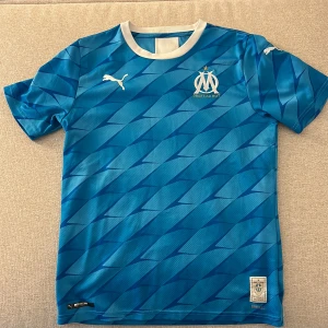 Blå Marseille fotbollströja Puma - Snygg blå fotbollströja från Puma med Olympique de Marseille-logga på bröstet och vit krage. Tröjan har ett randigt mönster i olika blå nyanser och korta ärmar. Tillverkad i lätt och ventilerande polyester, perfekt för träning eller match.