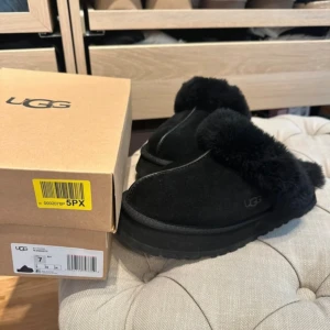 Svarta UGG slip-in med päls använda en gång!  - Mysiga slip-in skor från UGG i helsvart mocka med fluffig päls runt öppningen. Skorna har en platt sula med grovt mönster undertill och UGG-logga på sidan. Perfekta för chill dagar och håller fötterna varma med sin mjuka insida.
