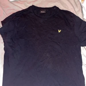 Svart t-shirt från Lyle & Scott XXL - Svart t-shirt från Lyle & Scott i storlek XXL. Klassisk rund hals och korta ärmar. På bröstet finns den ikoniska gula fågeln broderad. T-shirten är gjord i mjuk bomull och har en clean, stilren look som funkar till jeans eller shorts.