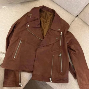 Brun skinnjacka från Filippa K - Snygg brun skinnjacka från Filippa K med klassisk bikerstil. Jackan har flera silvriga dragkedjor, bred krage och coola detaljer. Perfekt för dig som vill ha en tidlös och edgy look. Fodrad insida för extra komfort.