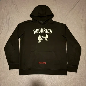 Svart Hoodie från Hoodrich  - Svart hoodie från Hoodrich med stor vit logga framtill och tryckta vita duvor. Baksidan har coolt motiv med duvor och röd text 'From Nothing To Something'. Huva med röd broderad text, känguruficka och mjukt material. Perfekt streetwear för dig som vill sticka ut.