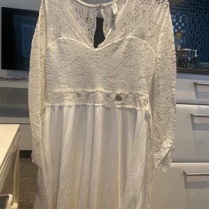 Vit långklänning med spets från Chi Chi London - Supersnygg vit långklänning från Chi Chi London med spets upptill och långa ärmar. Klänningen har v-ringad hals, spetsdetaljer och ett flowigt satintyg nedtill. Perfekt för dig som vill ha en elegant look med romantiska vibbar och fina detaljer.