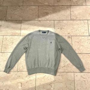 Grå stickad tröja från Polo Ralph Lauren - Snygg grå stickad tröja från Polo Ralph Lauren med klassisk rund hals och broderad logga på bröstet. Tröjan har ribbade muddar vid ärmslut och nederkant, och är långärmad. Perfekt för dig som gillar stilrena och tidlösa plagg. Om ni har några frågor är det bara att höra av sig. Skickar samma dag eller dagen därpå som senast!