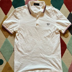 Vit Fred Perry pikétröja S - Vit pikétröja från Fred Perry i storlek S. Klassisk design med svart broderad logga på bröstet och krage med svarta och röda ränder. Tröjan har korta ärmar och är gjord i bomull med en snygg, normal passform. Perfekt för dig som gillar stilrena och sportiga plagg. Använd 1-3 gånger!