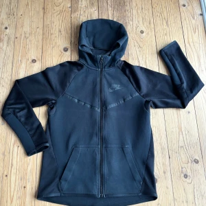 Svart hoodie från Nike  - Svart hoodie från Nike i storlek XL, passar dig som är 13-15 år. Tröjan har hel dragkedja, huva och två stora fickor framtill. Materialet är mjukt och har mesh-detaljer på ärmarna för extra ventilation. Snygg Nike-logga på bröstet.