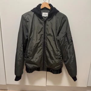 Grön höstjacka med huva från KappAhl (New York studios) - Snygg mörkgrön bomberjacka från KappAhl med svart huva och muddar. Jackan har dragkedja framtill, fickor på sidorna och är prydd med texten 'JUST CHILL' på mudden nedtill. Perfekt för dig som vill ha en cool och avslappnad stil.