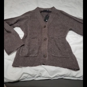 Brun stickad kofta med fickor - Mysig brun stickad kofta från Lager 157l med ribbade muddar och två stora fickor framtill. Koftan har v-ringad hals och stängs med tre knappar. Perfekt för lager-på-lager och chill dagar. Snygg och enkel att matcha med jeans eller leggings. I storlek M. Helt ny