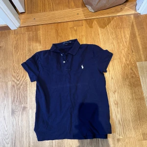 Mörkblå piké från Polo Ralph Lauren - Snygg mörkblå pikétröja från Polo Ralph Lauren i klassisk passform. Tröjan har korta ärmar, krage och två knappar framtill. Ikoniska Polo-loggan i vitt broderad på bröstet. Tillverkad i mjuk bomull som är skön mot huden.