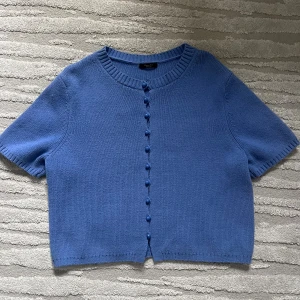 Blå kortärmad kofta Massimo Dutti - Kortärmad blå kofta från Massimo Dutti med rund hals och dekorativa knappar framtill. Stickad i mjukt material med ribbade detaljer vid hals och ärmslut. Perfekt för lager-på-lager och chill stil. Snygg att matcha med jeans eller kjol.