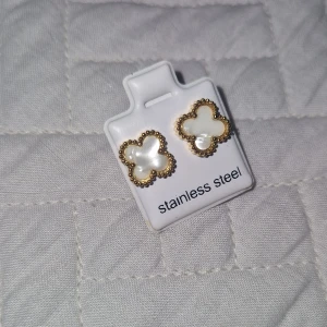 Guldiga fyrklöver örhängen i stål - Snygga örhängen i form av fyrklöver med pärlemorskimrande vit yta och guldfärgad kant. Tillverkade i rostfritt stål och har en elegant, tidlös design som passar perfekt till din stil. Helt nya, köpta i Spanien