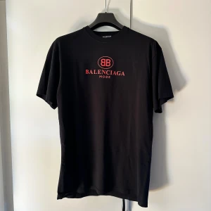 Svart Balenciaga t-shirt med röd logga - Svart t-shirt från Balenciaga med röd BB-logga och texten 'Balenciaga Mode' på bröstet. Klassisk rund hals och korta ärmar. Tillverkad i mjuk bomull för skön känsla. Perfekt för dig som gillar exklusiva streetwear-plagg.
