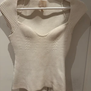 Ribbad vit stickad topp med v-ringning - Säljer en ribbad vit stickad topp med markerad v-ringning och breda axelband. Toppen har en figurnära passform och är ärmlös, perfekt att styla med jeans eller kjol. Materialet är mjukt och stretchigt vilket gör den superbekväm.