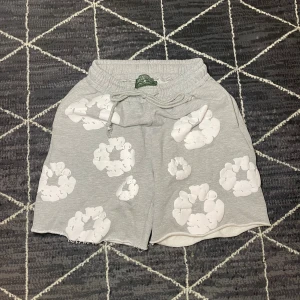 Grå shorts med vita moln från Denim Tears - Säljer ett par grå shorts från Denim Tears med vita molnliknande tryck över hela plagget. De har elastisk midja med snörning och är tillverkade i mjuk bomull. Perfekt för chill dagar och har en loose passform för extra komfort. storlek M