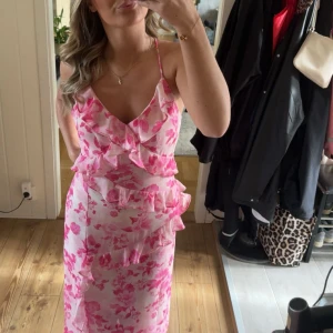 Rosa blommig långklänning med volanger - Superfin långklänning i ljusrosa och vitt med blommigt mönster och volangdetaljer. Klänningen har smala axelband, v-ringning och öppen rygg med knyt. Perfekt för sommarens och semestrarnas alla tillfällen när du vill sticka ut lite extra!!🩷Oanvänd och prislapp kvar
