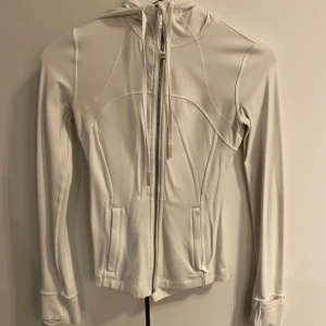 Vit hoodie från Lululemon med dragkedja - Supersnygg vit hoodie från Lululemon med figurnära passform och långa ärmar. Tröjan har huva med snörning, två fickor fram. INGA DEFFEKTER. Storleken är 2, vilket motsvarar en xs/s.