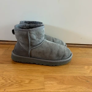 Grå UGG boots i mocka - Säljer ett par klassiska UGG boots i grå mocka med rund tå och platt sula. Skorna har en mjuk och fluffig insida som håller fötterna varma och bekväma. Perfekt för kyliga dagar och har UGG-logga baktill. Stilsäkra och tidlösa boots för dig som gillar komfort.