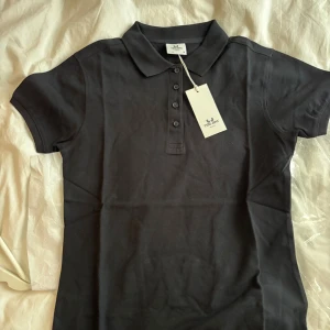 Svart pikétröja från Tee Jays, M - Klassisk svart pikétröja från Tee Jays med krage och knappar framtill! Tröjan är helt ny! Passar perfekt till exempelvis paddeln, tennisen eller golfen!🖤