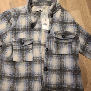 Rutig shirt från Only i blått och grått - Shirt från Only i mjukt material med rutigt mönster i blått, grått och vitt. Skjortan har klassisk krage, knäppning framtill och två bröstfickor med lock och knappar. Perfekt lager-på-lager-plagg för en chill look.