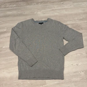 Grå stickad tröja Tommy Hilfiger - Snygg grå stickad tröja från Tommy Hilfiger med rund halsringning och diskret logga broderad på bröstet. Tröjan har ribbade muddar vid ärmslut och nederkant, perfekt för lager-på-lager-stil. Klassisk och stilren design som funkar till allt.