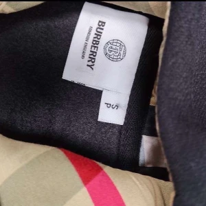 Svart hoodie med dragkedja - Svart hoodie från Burberry i storlek XS med klassisk beige-rutig huva och guldfärgad dragkedja. Hoodien har snörning vid huvan och är tillverkad i mjuk bomull. Perfekt för dig som vill ha en stilren och lyxig look med ikoniska Burberry-detaljer. 