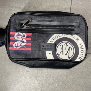 Svart axelväska med tryck - Snygg svart axelväska med coolt tryck och detaljer i vitt och rött. Väskan har en dragkedja framtill och ett mönster med ränder och grafiska element. Materialet är skinn och väskan har en modern, rektangulär form. Perfekt för dig som vill sticka ut.