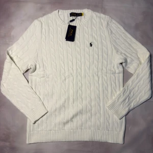 Vit kabelstickad tröja Polo Ralph Lauren - Vit kabelstickad tröja från Polo Ralph Lauren med klassisk rund hals och broderad logga på bröstet. Tröjan är tillverkad i mjuk bomull och har ribbade muddar vid ärmslut och nederkant. Perfekt för dig som gillar stilren och tidlös design.