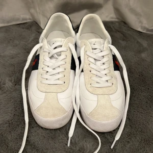 Vita sneakers från Polo Ralph Lauren - Snygga vita sneakers från Polo Ralph Lauren med marinblå detaljer och röd logga på sidan. Skorna har vit snörning, beige mockadetaljer och rund tå. Klassisk design med Polo-tryck på hälen och insidan. Perfekta för en stilren och sportig look.