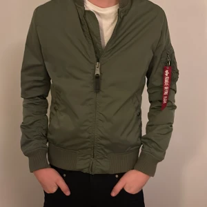 Grön bomberjacka från Alpha Industries - Säljer en snygg grön bomberjacka från Alpha Industries med klassisk röd 'Remove Before Flight'-tag på ärmen. Jackan har ribbade muddar, dragkedja framtill och fickor med knappstängning. Perfekt för dig som gillar streetwear och vill ha en ikonisk look. ALDRIG ANVÄND och PRIS KAN DISKUTERAS