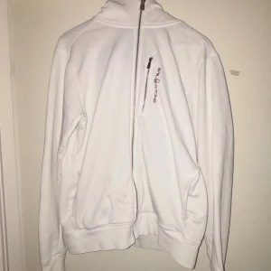 Vit ziptröja från Sail Racing - Vit långärmad ziptröja från Sail Racing med hög krage och dragkedja hela vägen framtill. Tröjan har en liten bröstficka med dragkedja och diskret logga på bröstet. Perfekt för dig som gillar stilren och sportig look.